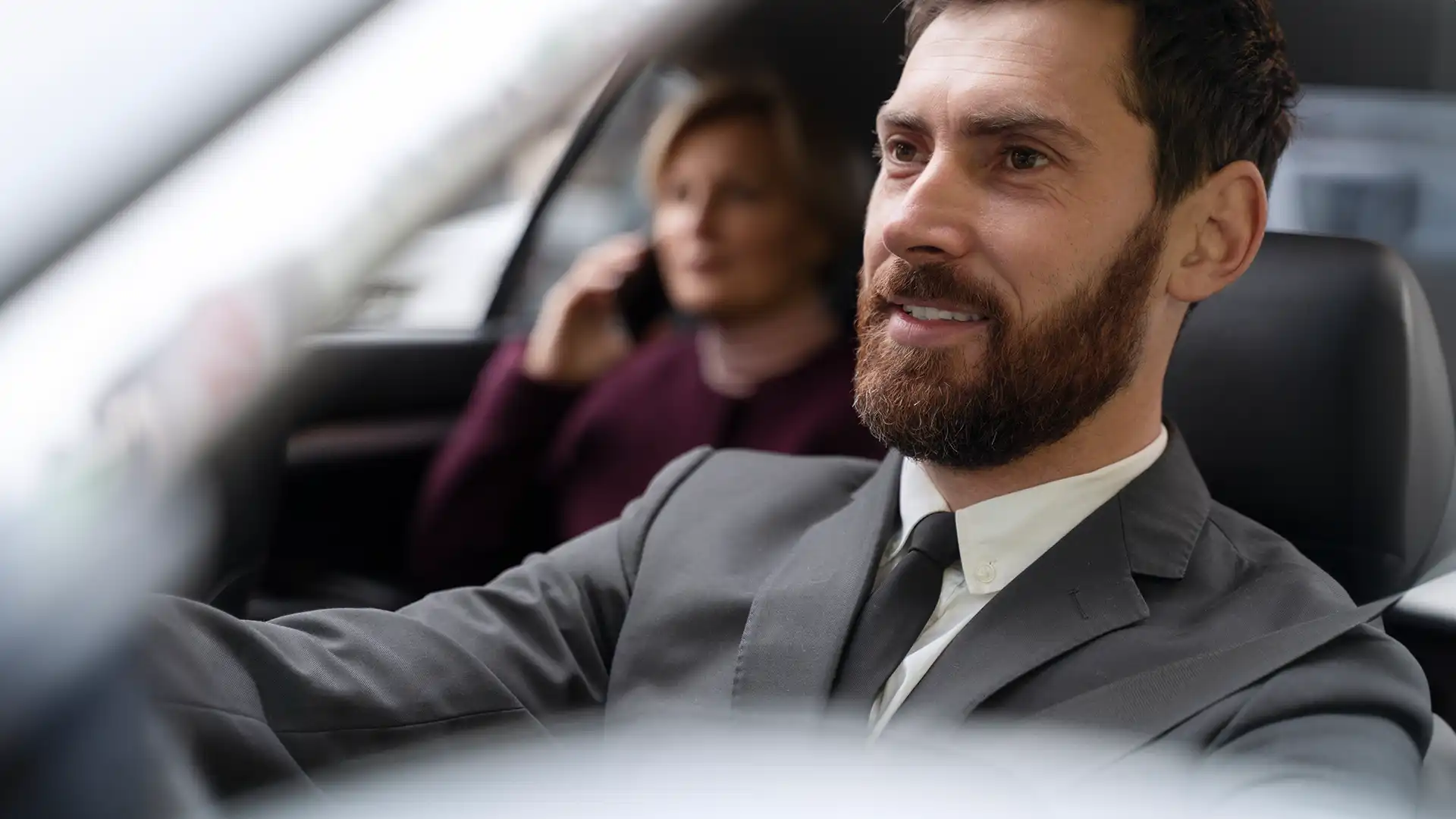 Foremost driver in a suit drives while smiling, with a woman on the phone in the backseat.