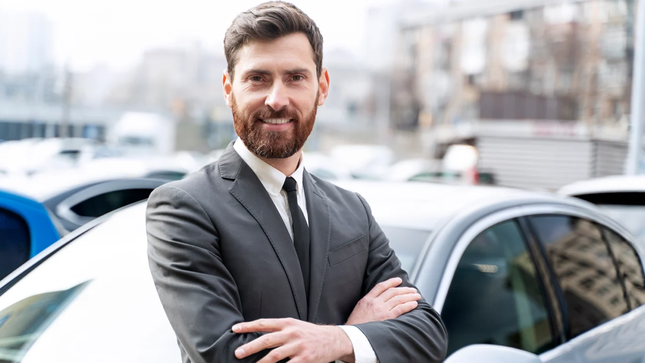 A man in a suit, arms crossed, smiles confidently in front of a car, showcasing personal driver services.