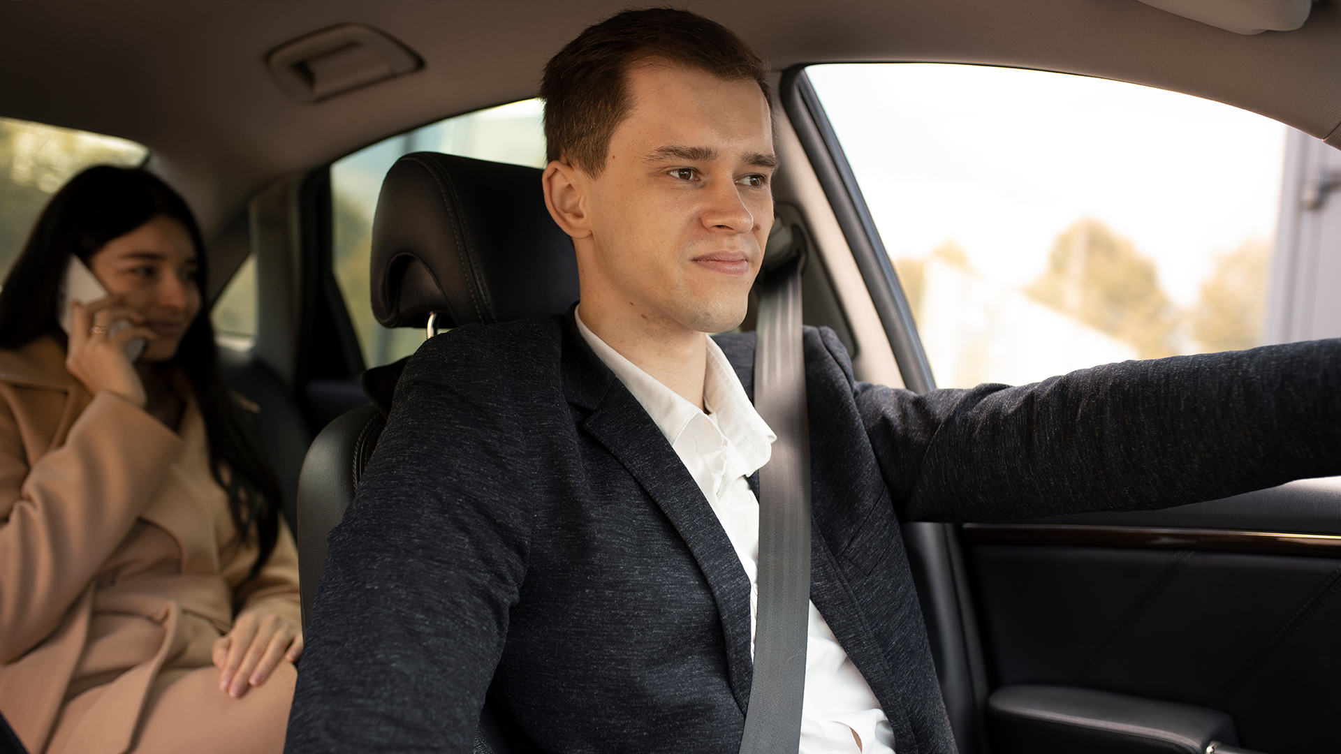 A personal driver in a white shirt and dark blazer driving a car, with a woman in the backseat talking on the phone.