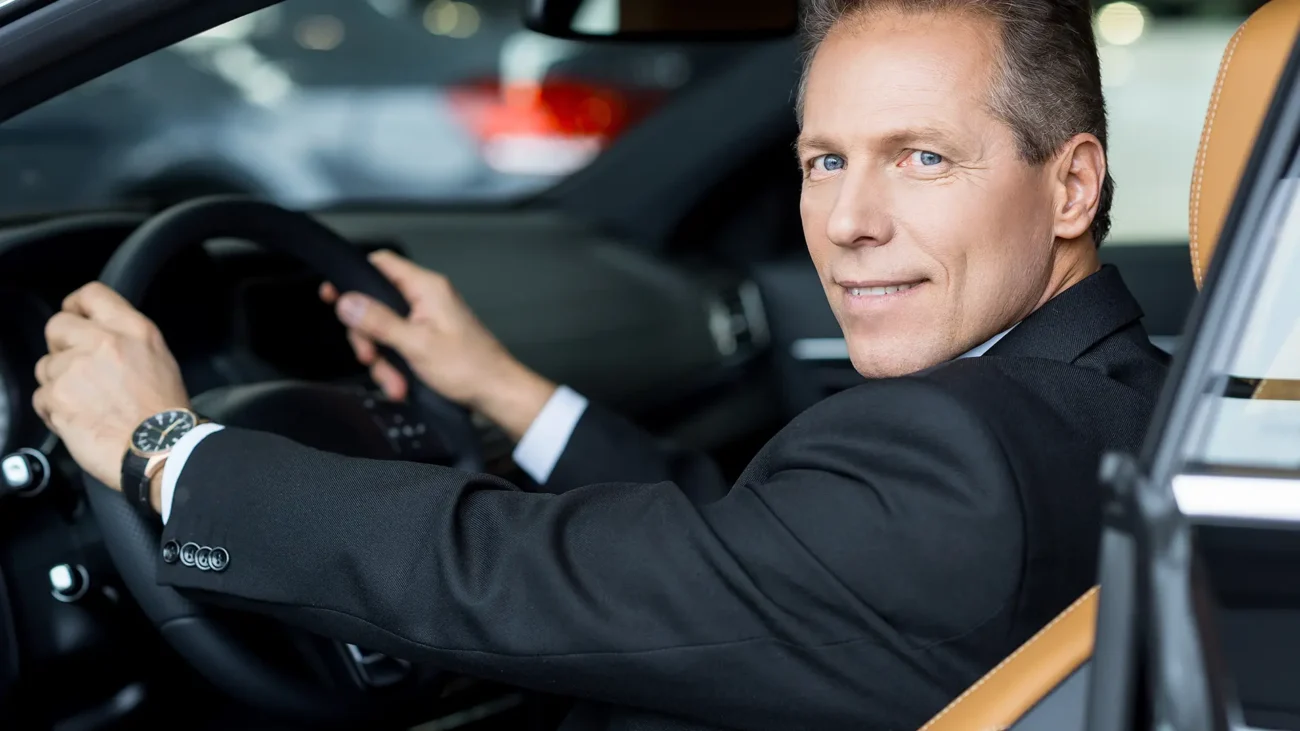 A professional chauffeur in a suit, smiling confidently while sitting in a car and holding the steering wheel. Safe driver services in Dubai.