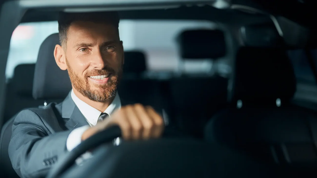 A professional chauffeur in Dubai, wearing a suit, is smiling while driving a luxury car.