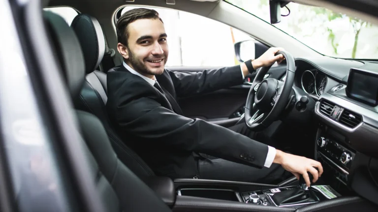 A smiling Event Driver-chauffeur in a black suit and tie sits in the driver's seat of a luxury car, ready to drive.