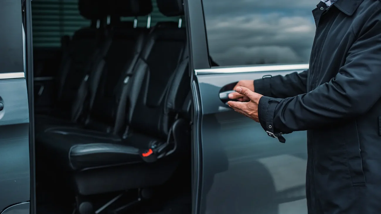 Event Driver in a dark jacket opens the side door of a van, revealing empty black leather seats inside.