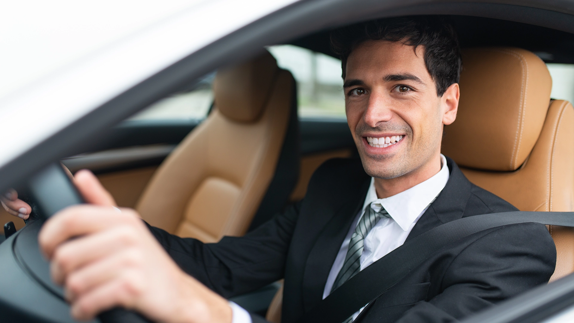 A professional chauffeur in a suit, smiling confidently while driving a car, representing Road Trip Driving Driver Service.