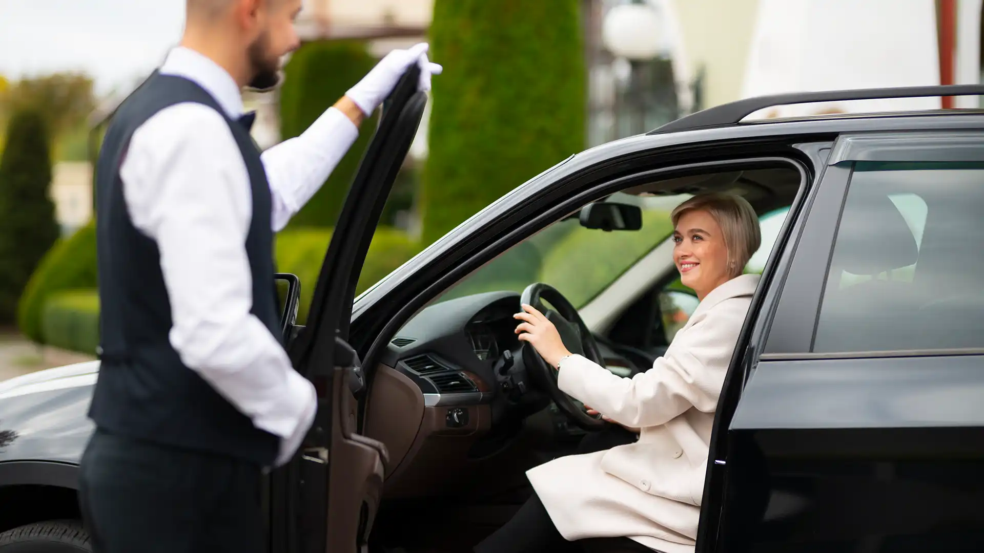 A valet opens the car door for a smiling woman in a white coat, highlighting premium Valet Parking Services.