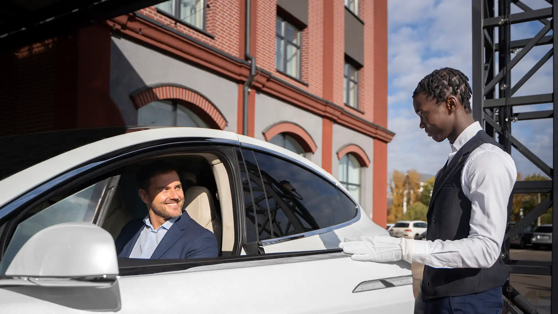 A valet in uniform assists a man in a suit sitting in his car, showcasing premium Valet Parking Services.