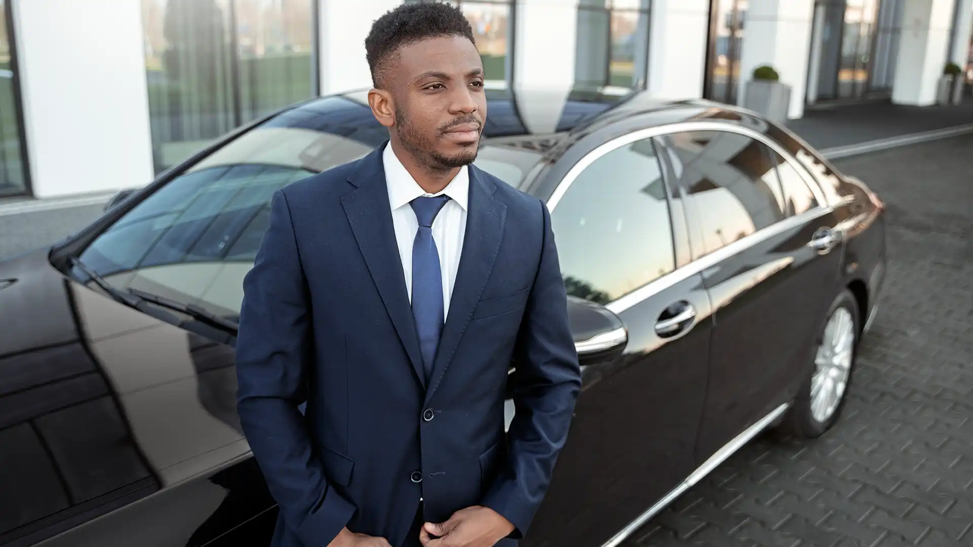 A professional chauffeur in a suit, standing confidently beside a black luxury car, representing City Tour Driving service.