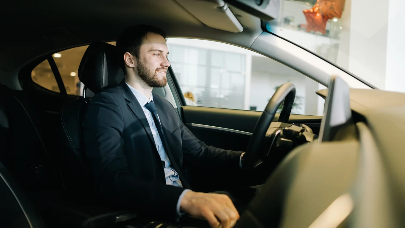 A well-dressed man, possibly a chauffeur, is seated in the driver's seat of a luxury car, smiling and appearing professional.