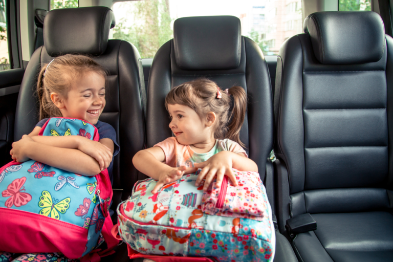 Two young girls with colorful backpacks sit happily in the backseat of a car, representing safe and cheerful school transport services.