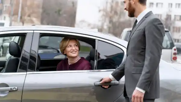 A professional chauffeur in a suit opens the car door for a smiling woman, exemplifying luxury airport chauffeur service.