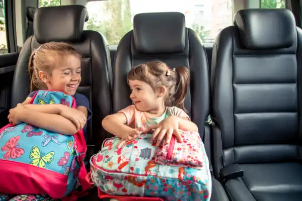 Two young girls with colorful backpacks smiling in a car's backseat, showcasing safe School Pick-Up and Drop-Off Services.